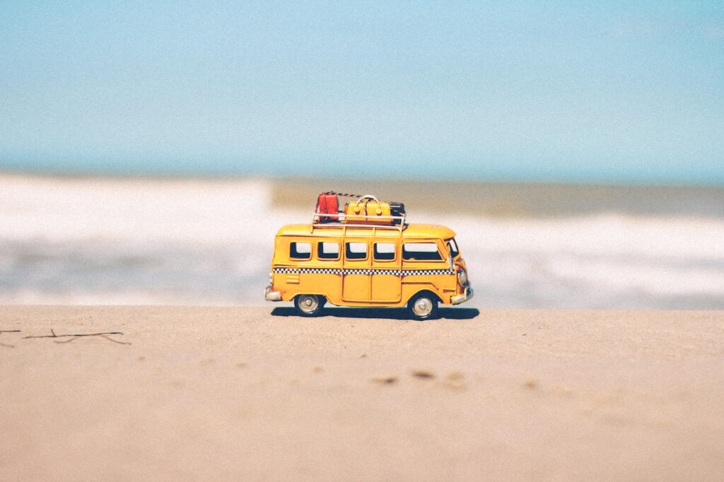 A Imagem tem uma van kombi de brinqueda na areia de uma praia com o mar de fundo desenfocado, para ilustrar o Transfer do Aeroporto de Porto Seguro Para Trancoso